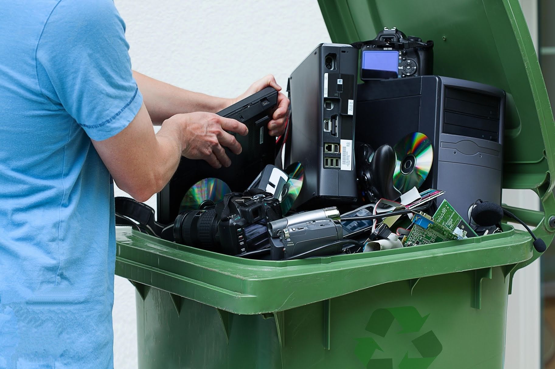 Electronics recycling disposal showing person discarding computer hardware, cameras, and electronic devices into green recycling bin with sustainability symbol